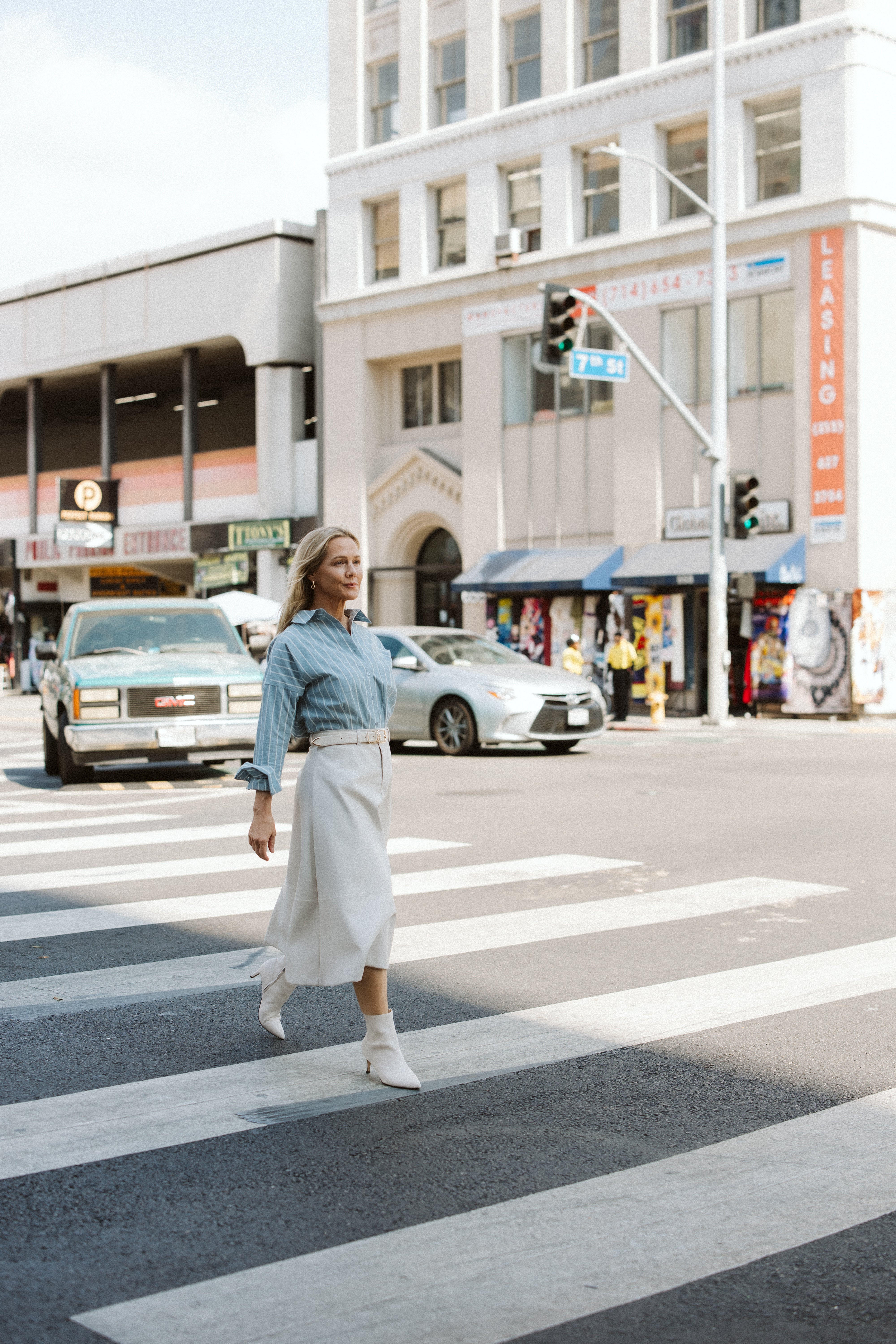 Jennie Garth crossing a New York street in a leather skirt and striped button down shirt in an urban setting with buildings and cars in the background